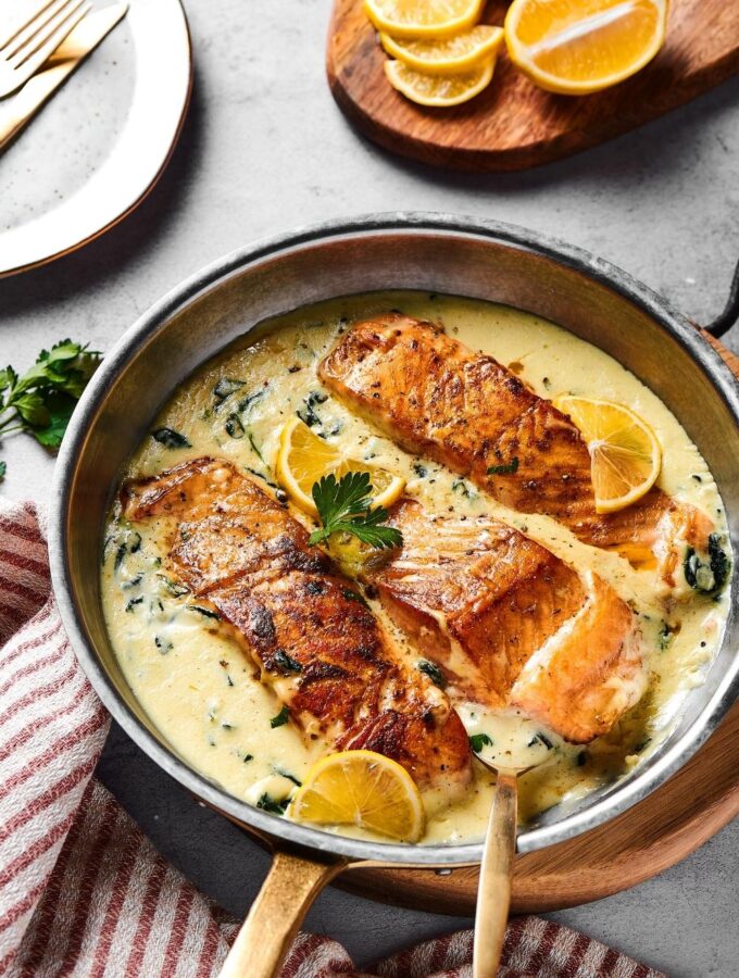 A skillet on a wood board filled with garlic butter sauce and three salmon fillets. Behind it is part of a wood cutting board with lemon wedges on it.