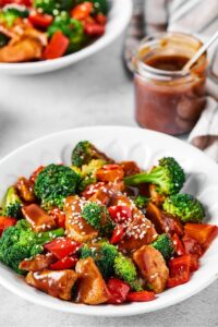 A white bowl with chicken stir fry in it. Behind it is a glass cup filled with stir fry sauce.