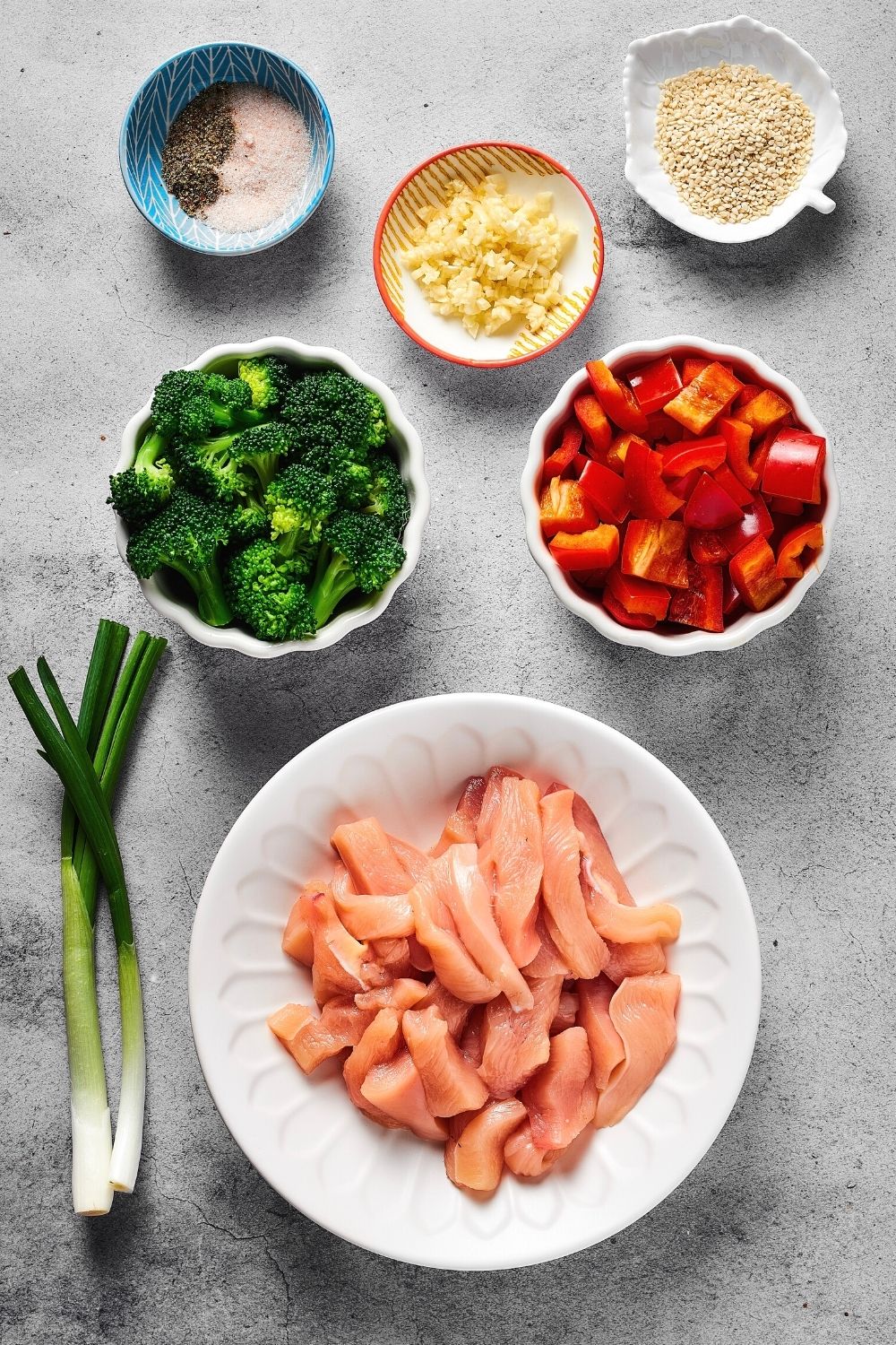 A white plate with chicken in it, a cup of broccoli, a cup of sliced red bell peppers, a plate with minced garlic on it, a bowl half filled with salt and half with pepper and a bowl of sesame seeds all on a grey counter.