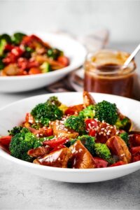 A white bowl that is filled with chicken stir fry. Behind it as part of a glass with stirfry sauce and part of another bowl with stir fry in it.