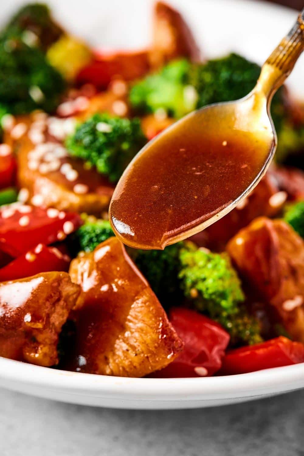 A spoon filled with stirfry sauce pouring it onto a piece of chicken. The chicken is on a plate with more chicken, broccoli, and red peppers.