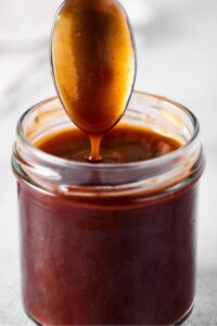 The head of a spoon with stirfry sauce on it dripping into stirfry sauce that is in a glass jar.