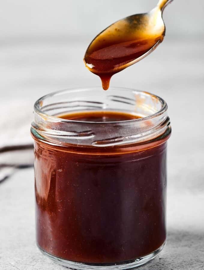 A glass jar filled with stirfry sauce. There's a spoon hovering over the jar with stirfry sauce on it and some dripping down from it.