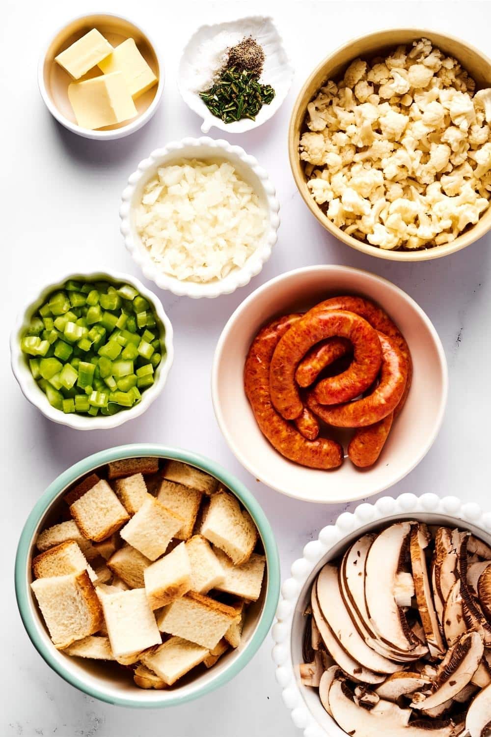 A bowl of bread slices, part of a bowl of mushrooms, a bowl of sausage, a small bowl of green peppers, a small bowl of onion, a small bowl of cauliflower, a small bowl of butter, and a small bowl of seasonings on the white counter.