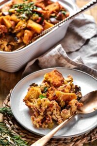 A white plate with stuffing on it with a spoon line next to it. Behind the plate is part of a white casserole dish that is filled with stuffing.