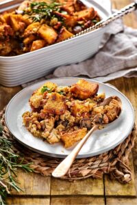 A white plate with stuffing on it and a spoon with stuffing on top and a straw placemat. Behind the plate is part of a casserole dish filled with stuffiung on a wood countertop.