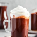 A glass mug that is filled with hot chocolate with whipped cream on top. The mugs on a gray played on a gray counter and in the background is part of a pitcher filled with hot chocolate.