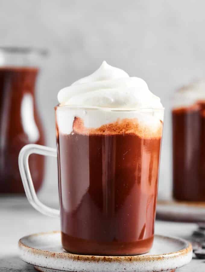 A glass mug that is filled with hot chocolate with whipped cream on top. The mugs on a gray played on a gray counter and in the background is part of a pitcher filled with hot chocolate.