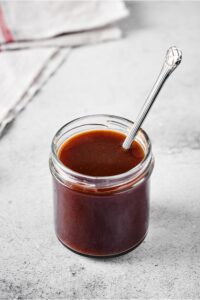 A glass jar filled with stirfry sauce with a utensil submerged in it. The glass jar is on top of a gray counter.