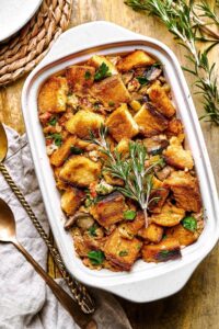 A white casserole dish filled with stuffing on top of the wooden counter. It's a left on the dish is part of two spoons and a gray napkin.