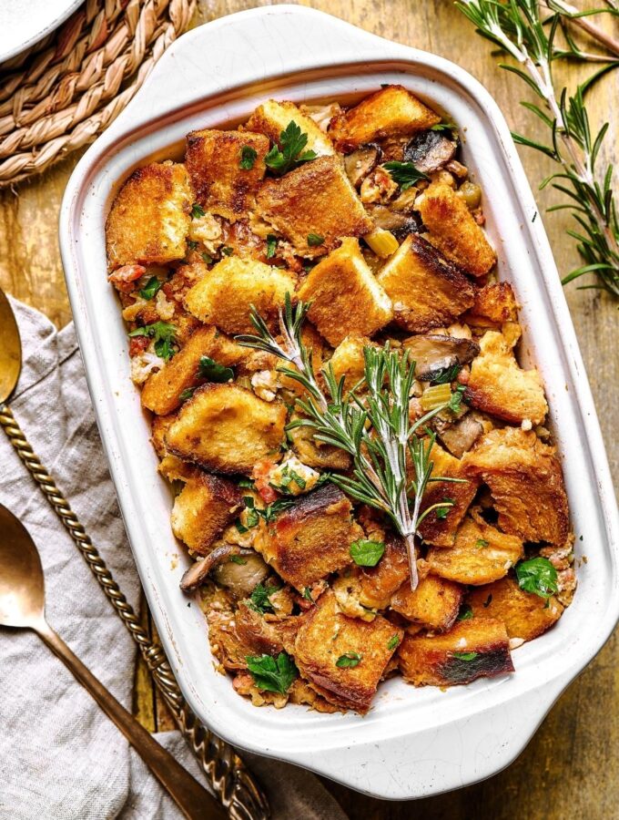 A white casserole dish filled with stuffing on top of the wooden counter. It's a left on the dish is part of two spoons and a gray napkin.