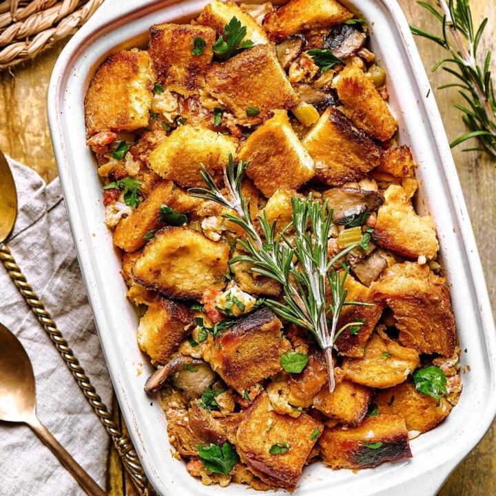 A white casserole dish filled with stuffing on top of the wooden counter. It's a left on the dish is part of two spoons and a gray napkin.