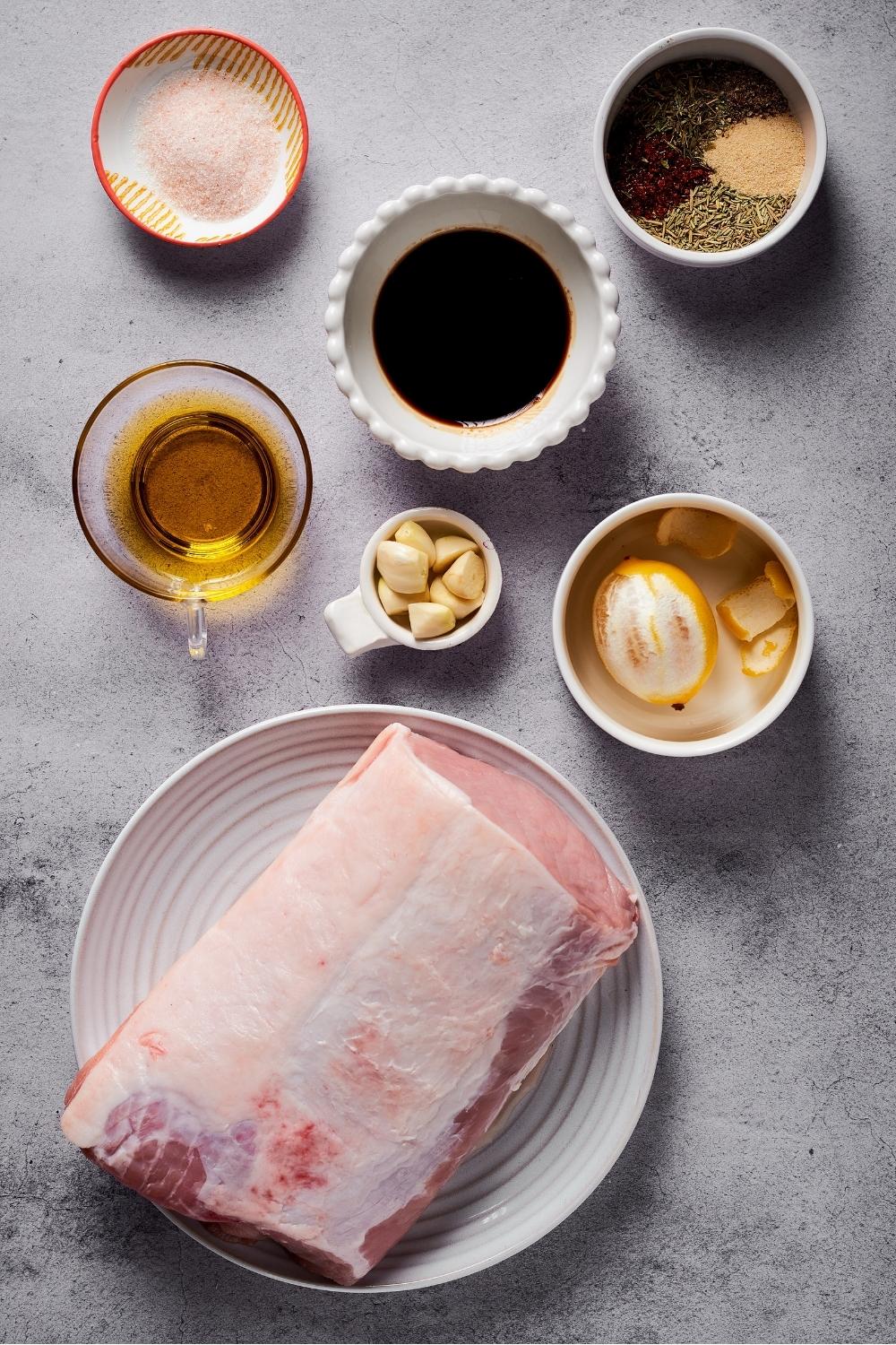 A white plate with a pork loin on it, a cup of lemon, a cup of balsamic vinegar, a bowl of seasonings, and a pitcher of olive oil all on a grey counter.