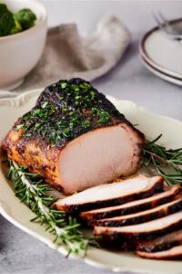 A white plate placed diagonally with a pork loin on it and a bunch of slices of the pork loin overlapping one another in front of it.