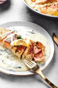 Part of a plate with part of a salmon fillet on it and a garlic butter sauce. There's a fork at the front of the fillet and some pieces cut out of the fillet leaned in front of it.