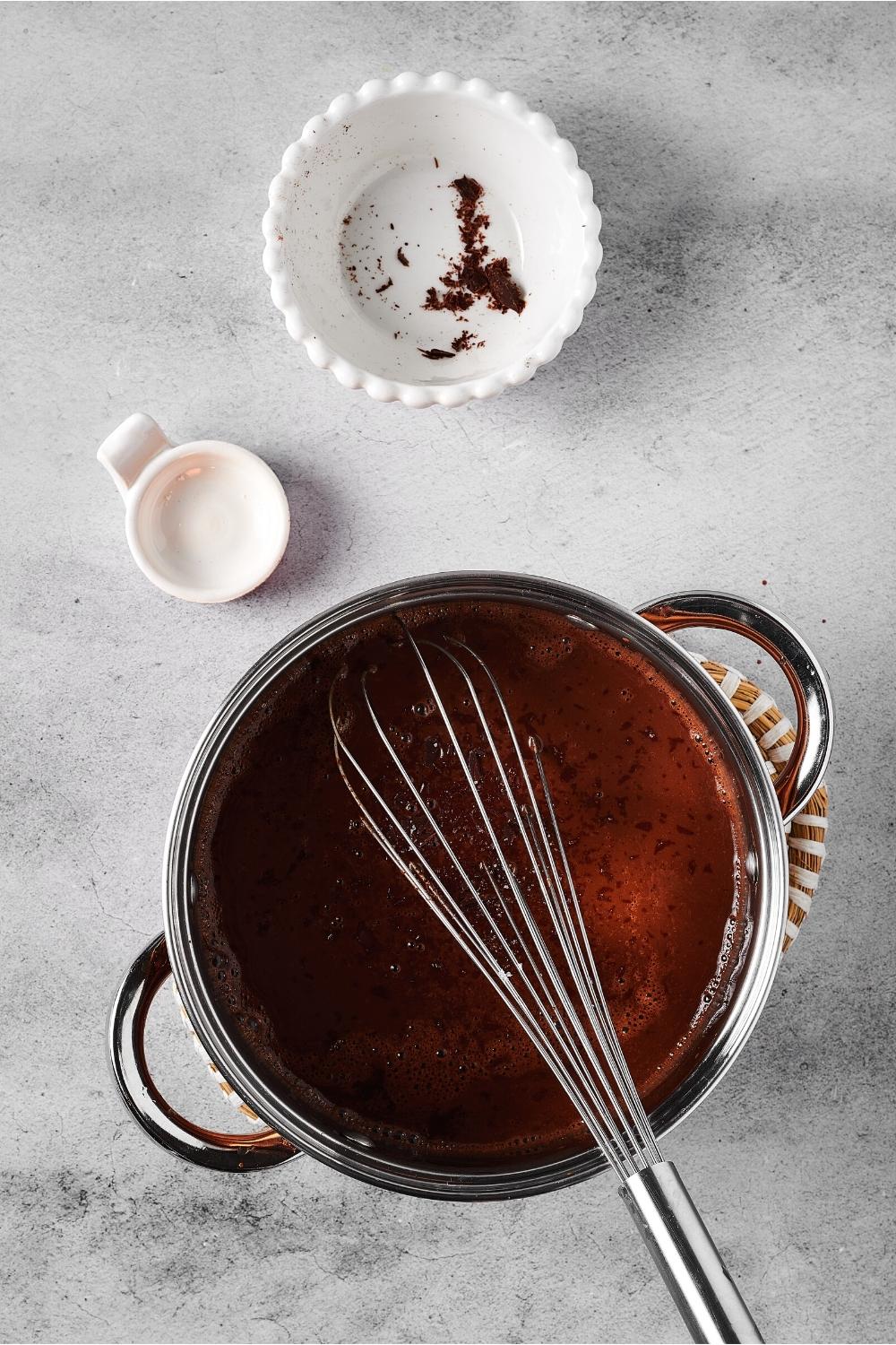 A pot filled with hot chocolate with a whisk in it. Behind it is two empty white bowls on a gray counter.