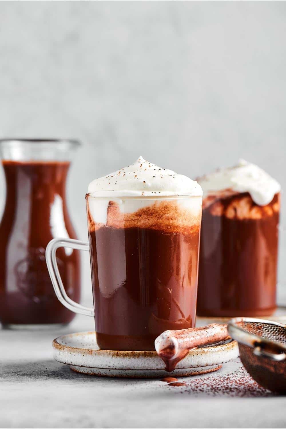 A glass cup of hot chocolate with whipped cream on top and a small plate. Behind it to the right is another cup of hot chocolate and to the left of that as a glass pitcher filled with hot chocolate.