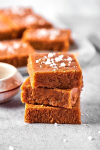 Three squares of peanut butter fudge sat on top of one another on a gray counter. In the background are other squares of peanut butter fudge.