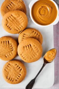 Part of a whiteboard with six peanut butter cookies on it along with a white cup of peanut butter and a spoon with peanut butter.