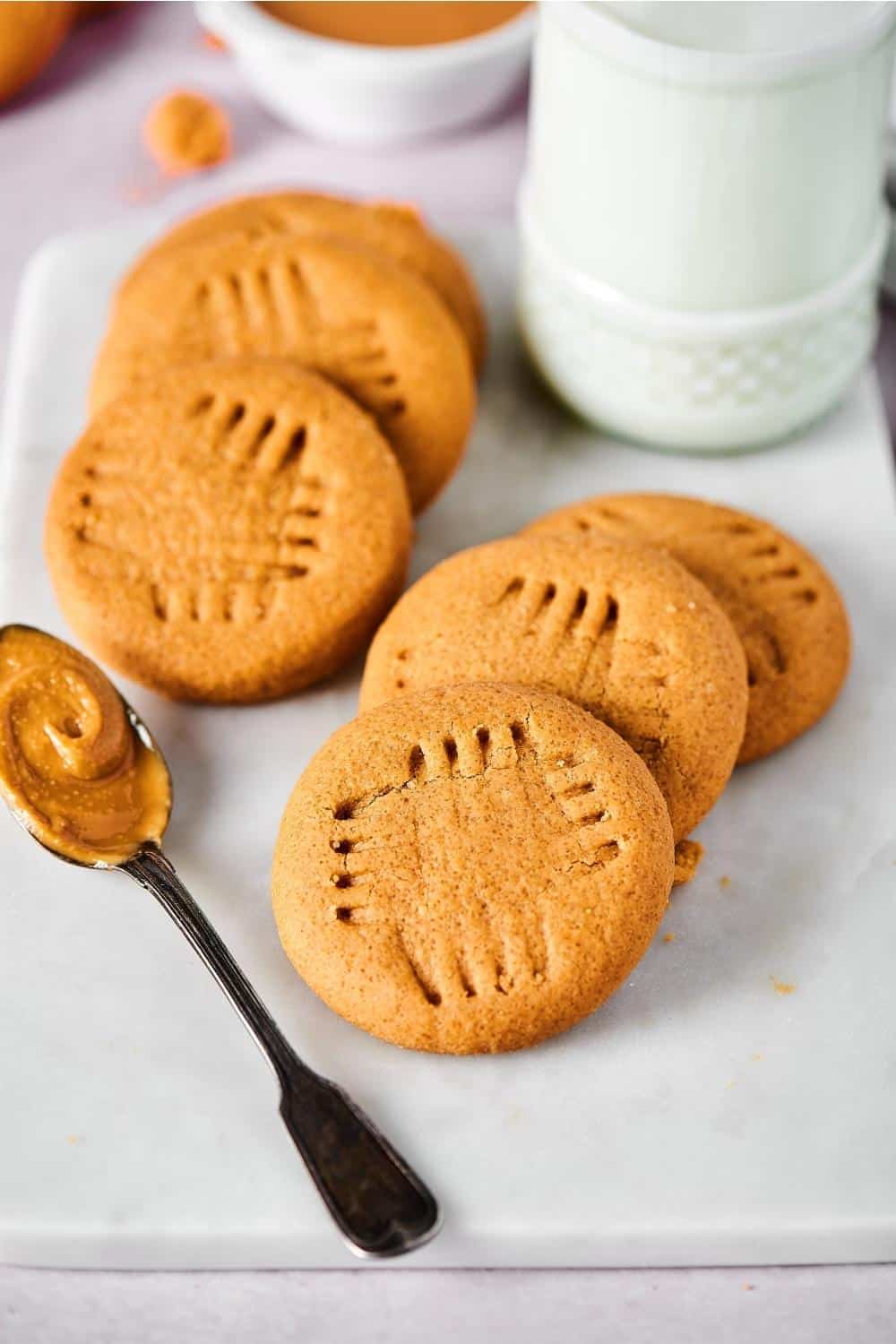 Three peanut butter cookies overlapping one another with three more peanut butter cookies overlapping one another to left on them. The cookies are on a whiteboard with a spoon with peanut butter on it in front of them.