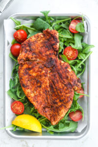 A cooked chicken breast on a bed of greens with tomatoes and a lemon wedge.