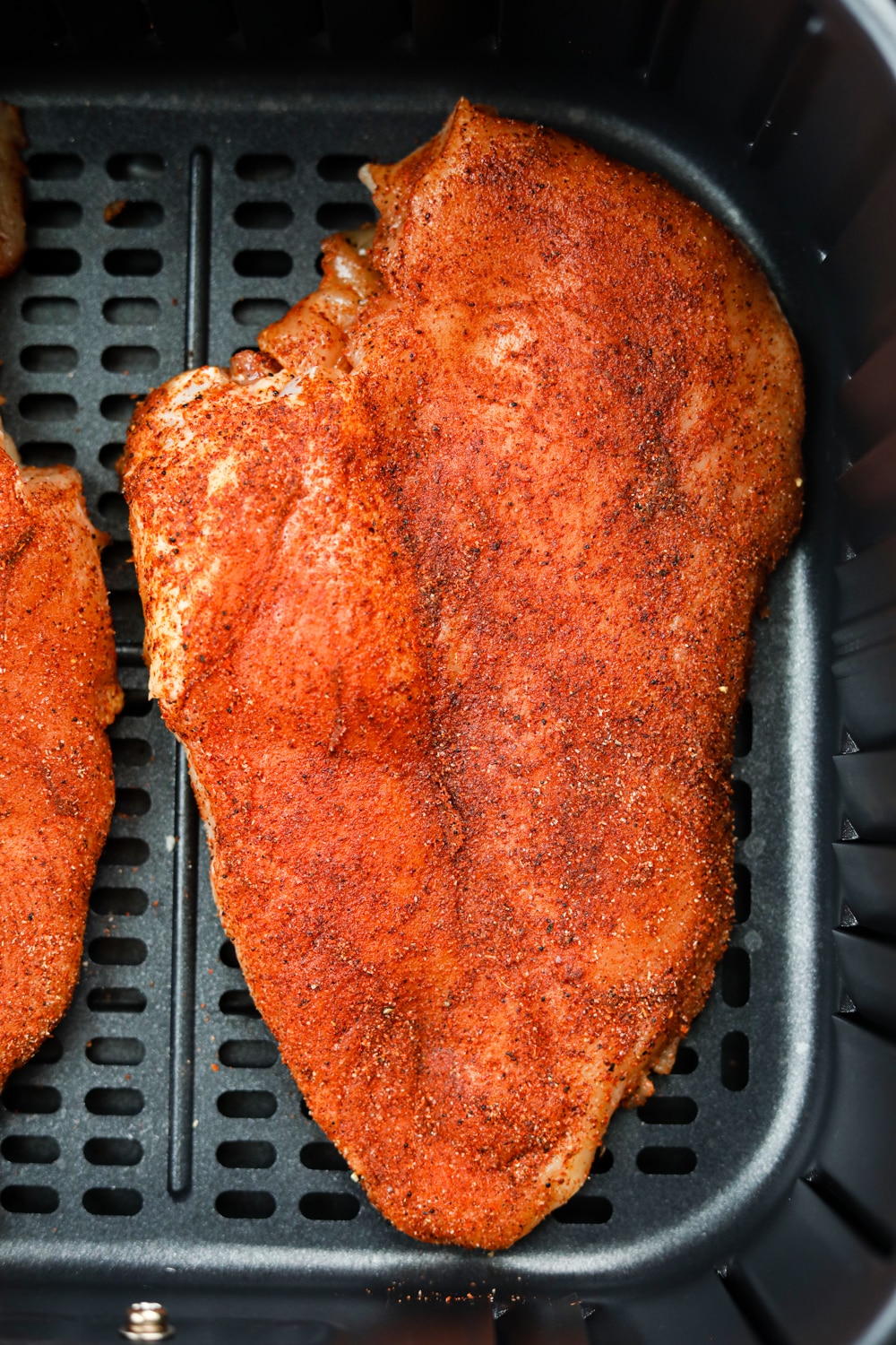 An uncooked chicken breast covered in a dry rub. The chicken breast is in an air fryer basket.