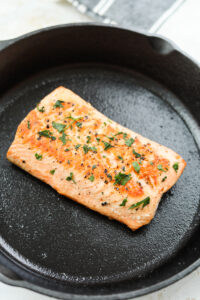 A salmon filet topped with salt, pepper, and fresh parsley in a black cast iron pan.