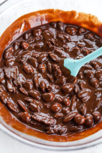 A glass bowl filled with mixed nuts that have been covered in melted chocolate. There is a blue spatula in the bowl.