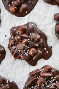 Chocolate nut clusters on a sheet of white parchment paper. The nut clusters are topped with flakey salt.