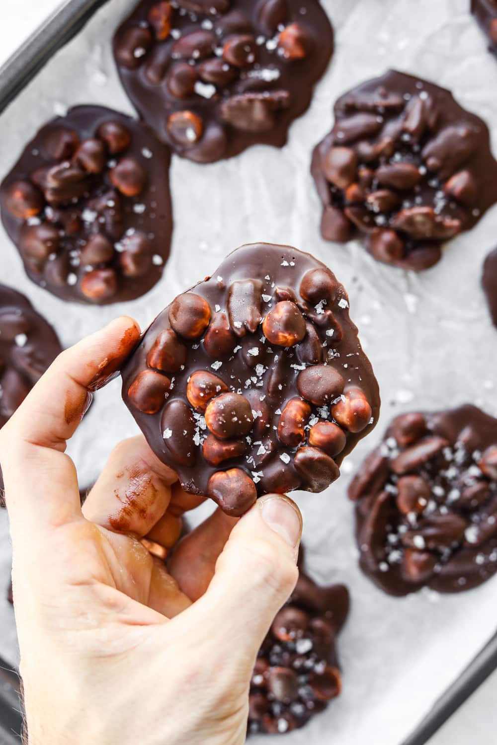 A hand holding a chocolate nut cluster. There's a tray of nut clusters underneath the hand.