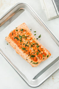 A filet of cooked salmon on a sheet of parchment paper set inside a tin tray. The salmon is topped with fresh parsley.