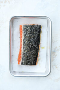 A filet of salmon faced skin side up in a tin tray. The salmon is coated in olive oil, salt, and pepper.