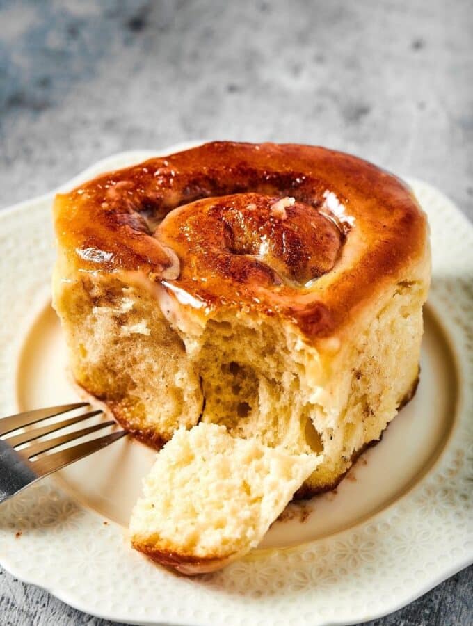 A cinnamon roll on a white plate. There is the head of a fork on the plate with a piece from the front of the cinnamon roll next to the fork.