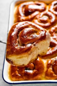 Hey cinnamon roll being held by utensil in front of a baking dish filled with the cinnamon rolls.
