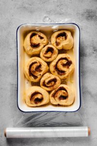 A baking dish with eight uncooked cinnamon rolls in it with plastic wrap sealed on top.