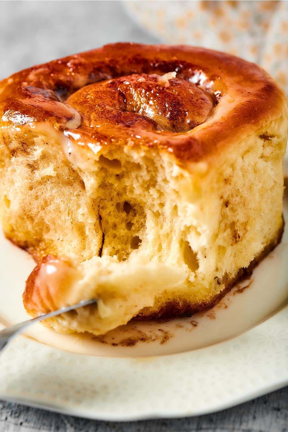 A cinnamon roll on a white plate. A fork is pulling away a piece form the front of the cinnamon roll.