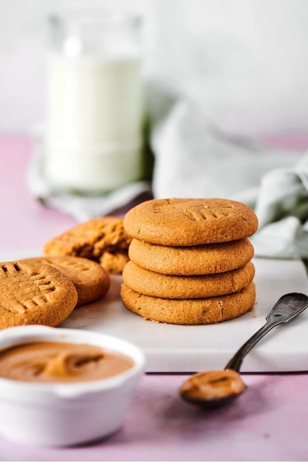 A white bowl filled with peanut butter in front of a white cutting board that has a stack of four peanut butter cookies on it with a few more cookies to the left of the stack.