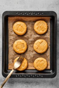 Two rolls of three peanut butter cookies on top of a piece of parchment paper on a baking sheet. There is a crisscross pattern in the cookies and a fork is on top of one of the cookies.