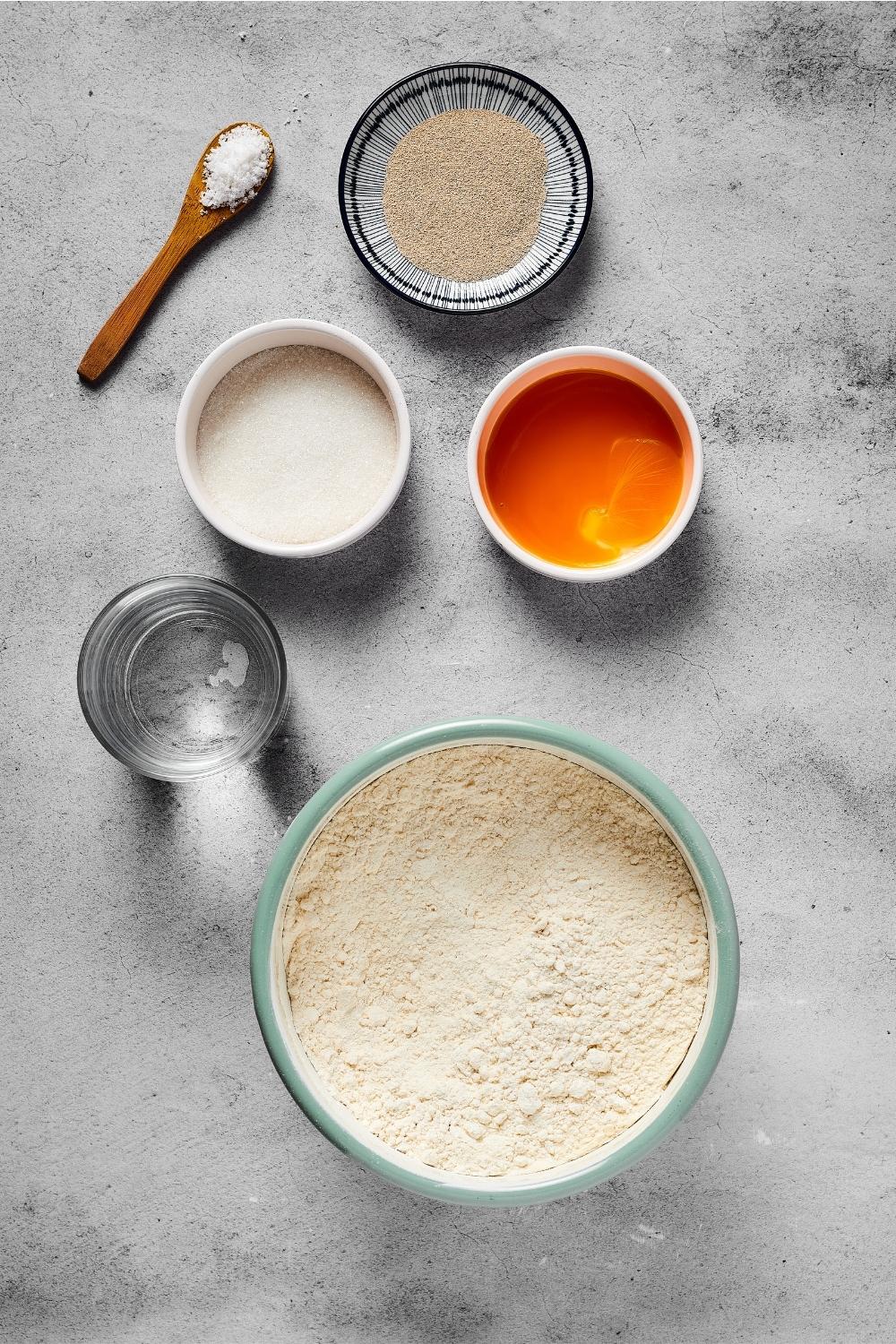 A bowl of flour, small white bowl of egg yolk, small white bowl of sugar, a small plate of yeast, and a small spoon with salt on it all on a gray counter.