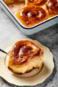 A cinnamon roll on a white plate with part of a baking tray with the cinnamon rolls in it behind it.