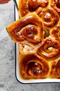 A cinnamon roll being held with a utensil over a baking dish of cinnamon rolls.