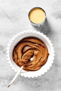 A white bowl filled with peanut butter with a spoon in it and a can of sweetened condensed milk behind it both on our gray counter.