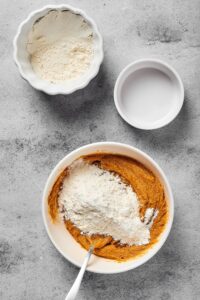 A white bowl filled with peanut butter with flour on top, an empty white bowl behind it, and a bowl with some flour in it all on a gray counter.