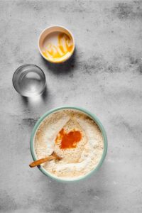 A bowl with flour, sugar, a girl, and water in it with a wooden spoon submerged in the bowl. Behind it is an empty cup of water in an empty white bowl that had the egg yolk in it all on a gray counter.