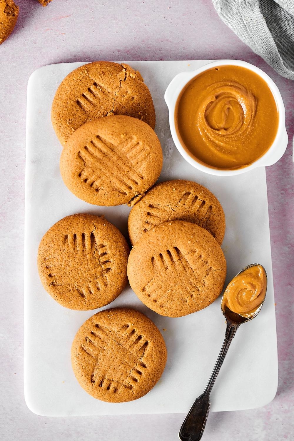 Six peanut butter cookies on a white cutting board with a white bowl filled with peanut butter on it and a spoon with peanut butter on it.
