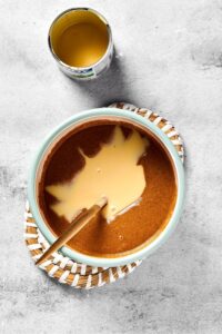 Melted peanut butter and sweeten condensed milk in a bowl and a straw placemat on a gray counter. There is a spoon in the bowl and an empty can of sweetened condensed milk behind the bowl.