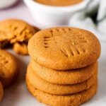 Hey stack of four peanut butter cookies and a white surface. Behind the stack is white bowl of peanut butter.