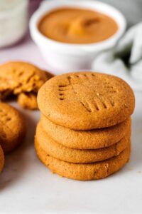 Hey stack of four peanut butter cookies and a white surface. Behind the stack is white bowl of peanut butter.