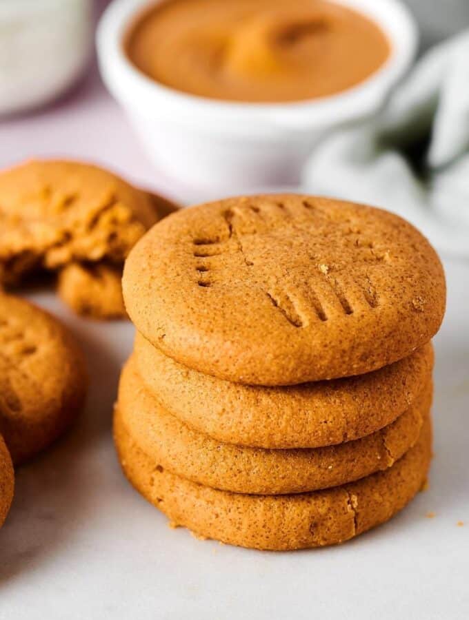 Hey stack of four peanut butter cookies and a white surface. Behind the stack is white bowl of peanut butter.