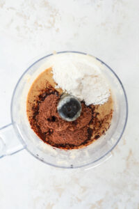 A food processor full of hazelnut butter, sweetener, vanilla extract, and cocoa powder.